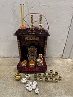 Full view of wooden temple shrine with figurines, incense holders, brass cups, incense sticks, and paper-wrapped items on floor in front.