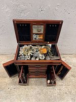 Photo showing open wooden jewelry box filled with assorted jewelry including necklaces, brooches, bracelets and rings, with compartments and drawers open.