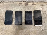 Four old smartphones shown side by side on a rough surface. Samsung, Motorola, Sony, and LG brands visible. Screens black and worn, one screen cracked.