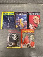 Five Astounding Science Fiction paperback books arranged on a metal surface showing colorful sci-fi themed covers including characters and machinery typical of 1950s sci-fi pulp art.