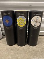 Three black cylindrical containers for Ritzenhoff beer glasses with different artistic labels shown side-by-side