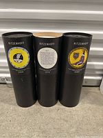 Three black cylindrical tubes labeled Ritzenhoff placed upright, showing the labels on each tube with colorful design artwork and edition numbers.