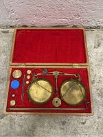 Photo of the brass balance scale set with its components positioned inside the wooden velvet-lined case, showing the balance beam, pans, weights, tweezers, rod, and small blue container.