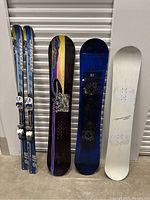 Full view of 2 Head skis and 3 snowboards lined up upright showing bindings and graphics