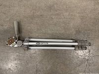 Folded view of silver Optex DPT175 tripod including the head and handle on concrete floor.