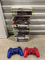 Stack of 27 Xbox 360 and PS3 game cases with two PS3 wireless controllers in front (blue and red)