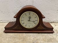 Full view of the wooden mantel clock with arched top and flat base placed on floor against white wall.