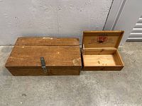 Two wooden crates placed on concrete floor, one larger with crest logo on top and one smaller open box with crest inside lid and metal clasp closing mechanism.