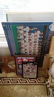 Photo showing stack of baseball memorabilia leaning against wall including framed pictures and large poster, in a living room area