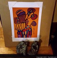 Full view of unframed Norval Morrisseau print attached to cardboard with two dark carved figurines in the foreground