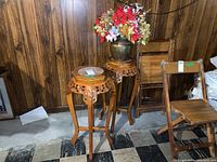 Two wood carved plant stands with marble tops beside a wooden folding chair on black and white tiled floor with wood panel wall background. Christmas floral arrangement on one table.