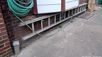 Aluminum extension ladder resting horizontally along brick wall outside, close to garden hose and concrete ground.