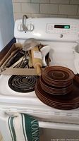 Stacked brown Wedgwood plates and bowl-shaped items on stove next to box of kitchen utensils including wooden rolling pin, large spoon, and spatulas.