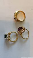 Photo showing three rings: one gold with missing stone and two 18k gold plated with green and purple stones.
