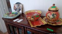 Overview of lot showing various rooster-themed ceramic dishes and plates arranged on wooden furniture.