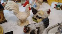 Three rooster and hen figurines on table with other household items. Tall white rooster stands prominently with a strong tail and comb.