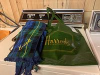 Blue and green tartan scarf folded next to and partially inside a green Harrods branded reusable tote bag on top of a washing machine.