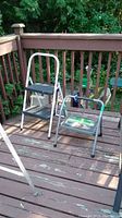 Two step stools on a wooden deck: one tall white and black metal and plastic step stool and a shorter gray metal and plastic step stool with padded handle showing wear