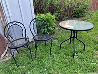 Full view of metal glass bistro set featuring two matching chairs with mesh seats and backs and a round glass-top table on grass.