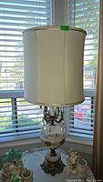 Full view of the vintage table lamp including the fabric shade, hand-painted glass body, and lower base seen in front of a window.