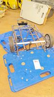 Metal collapsible hand truck resting on one of the blue plastic rolling dollies, showing wheels, folding frame, and handle.