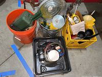 Wide view showing orange bucket with tools, yellow crate with funnels and scoops, galvanized pail, and black basin with pump devices.