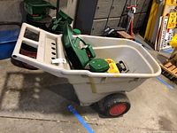White Rubbermaid garden cart containing a green hand held spreader and a yellow insect control sprayer, viewed from an angle showing the side and handle of the cart.