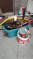 Green plastic bin filled with assorted hand tools including hammers, axes, crowbar, and saws, alongside a white bucket with smaller tools and parts.