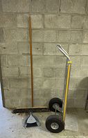 Full view of the hand truck, broom, dustpan, and brush against a concrete block wall.