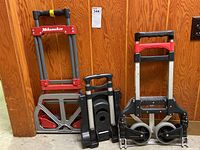 Three foldable hand trucks positioned upright against a wooden paneled wall, one Milwaukee brand with red and gray frame visible.