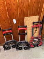 Photo showing all three foldable hand trucks standing upright against a wooden wall. Two are black and red with large dual wheels, and one is red with smaller wheels and elastic securing straps.