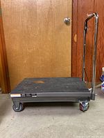 Side view of the metal lifting moving trolley showing the rubber mat surface, four wheels including one with a red brake mechanism, and the chrome handle with foot pedal.