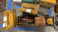 Three yellow metal military boxes showing visible wear and containing assorted tools including pliers, screwdrivers, and chains, alongside a brown leather tool holster.