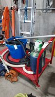 Full view of red flat bed trolley with blue plastic bucket, vacuum-like hoses, and a metal container on it, captured in industrial setting.