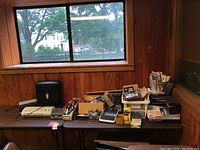 Wide photo showing a long table against wooden walls with various office supplies including a shredder, laminator, stapler, boxes and baskets with pens, pencils, calculators, markers and envelopes.