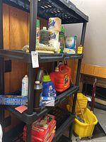 Front view of black plastic shelving unit with various household items stored on shelves. Shelving has vertical posts and slatted shelves.