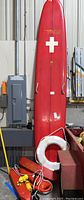 Full view of the large red rescue board standing upright against a wall with white cross symbol. Orange lifeguard belts and white lifebuoy ring leaning against the base of the board.