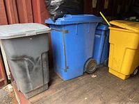 Four lidded trash bins in grey, blue, yellow colors, positioned side by side showing mobility wheels and general condition.