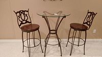 Full view of metal bar table with glass top and two matching metal stools with wooden seats and decorative backs