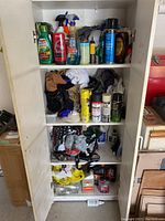 Four-shelf white cabinet full of car detailing supplies, spray paint, rags, gloves, various automotive fluids, and a hanging light.