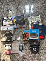 Photo of various Harley Davidson and motorcycle-related books and magazines with motorcycle bags arranged on a floor with a stone tile pattern.