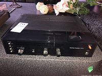 Front angle view of Harman Kardon 230e receiver showing top with ventilation slots and front controls with function selector and knobs.