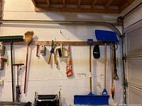 Photo showing hanging garden tools including broom, dust pan, small saws, hedge clippers, blue snow shovels on garage wall.
