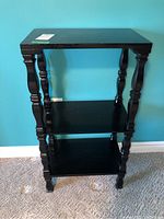 Full front view of black wooden shelving unit showing three flat shelves and decorative turned legs against blue wall and carpeted floor.