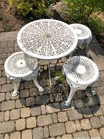 Full set of white cast iron outdoor round table and three stools on a stone patio with visible weathering and leaf debris.