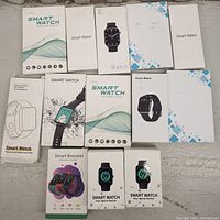 Lot of 13 smart watches and smart bracelets in original packaging arranged on a flat surface.
