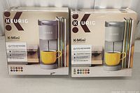 Two factory boxed Keurig K-Mini single serve coffee makers in studio gray color with yellow cup shown on packaging.