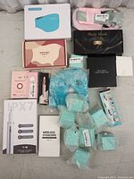 Lot with blue face mask box, pink warm belt box, black box with intelligent pose corrector, blue gel eye mask, wireless otoscope box, ultrasonic dental scaler box, pink nano mist sprayer box, and several bubble wrapped items.