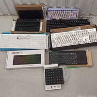 Photo showing multiple keyboards including white wired keyboards, black wireless keyboard with folio, colorful round keycap keyboard, HK Gaming mechanical keyboard in packaging, black numeric keypad, and more.
