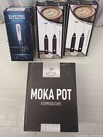 Photo shows three boxed items including two identical black electric milk frothers and one boxed electric white milk frother and one boxed Moka pot.